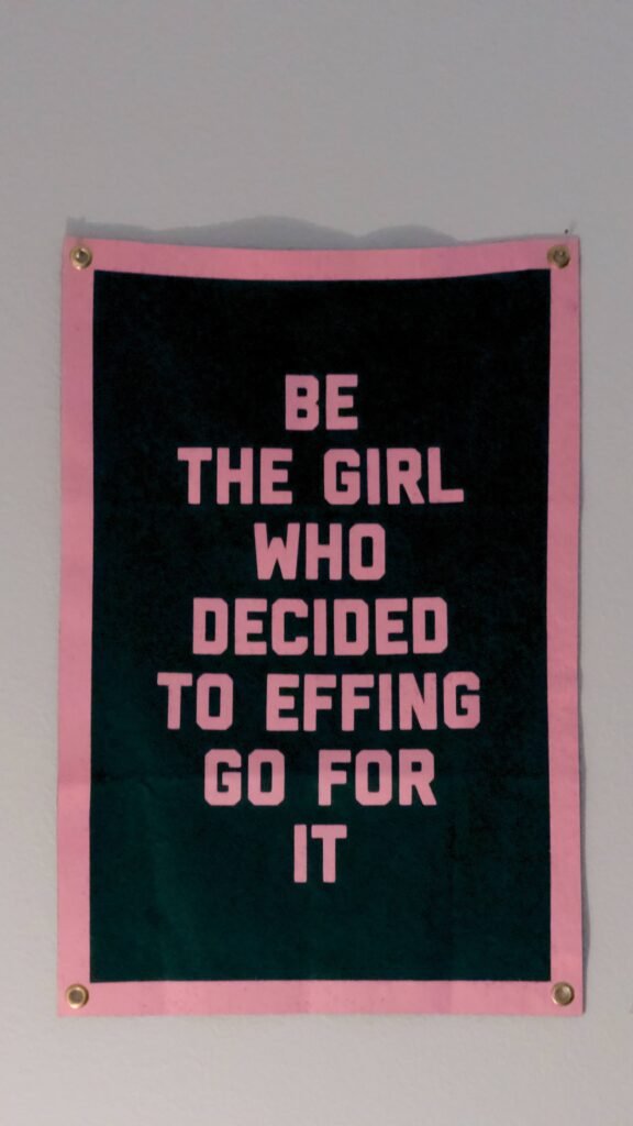 A black and pink poster on a wall saying "Be the girl who decided to effing go for it".