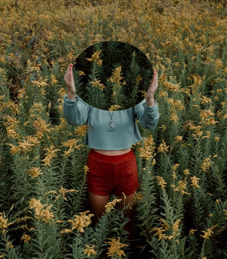 A woman standing in the middle of a flower field, holding a mirror in front of her head.