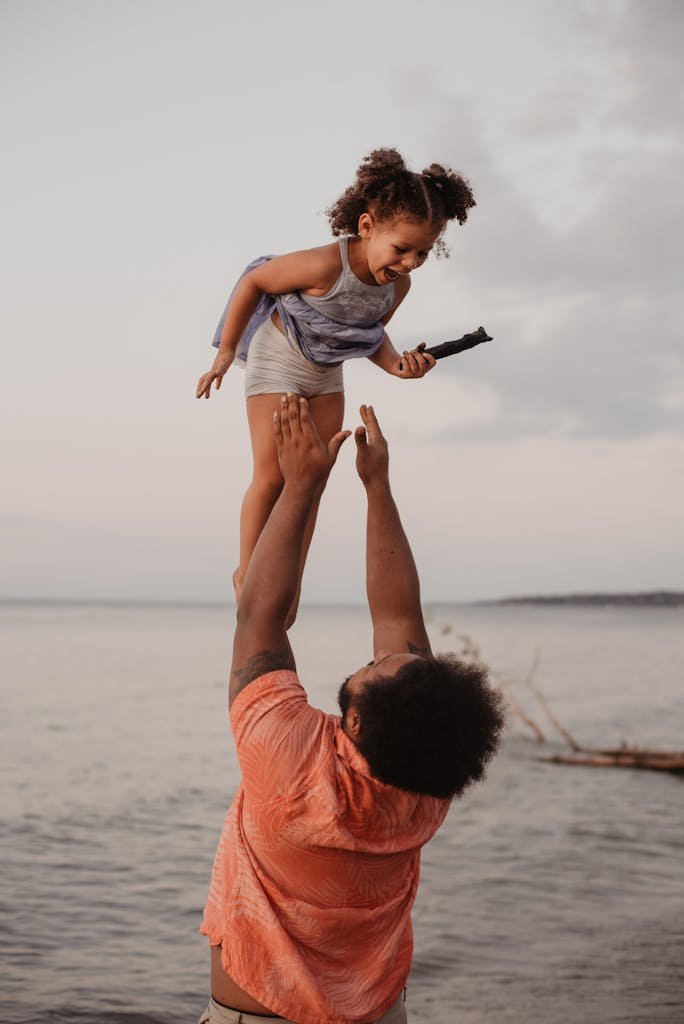 A joyful father lifts his daughter in the air by the beach during sunset, capturing a moment of love and happiness.